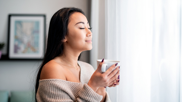 young peaceful woman holding cup of tea next to window