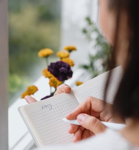 over shoulder view of girl writing in diary next to windowsill with yellow flowers